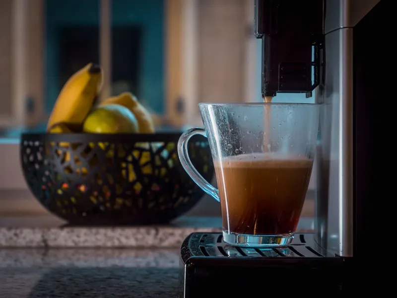 Glass cup catching freshly brewed coffee near a bowl of bananas and apples in a modern kitchen