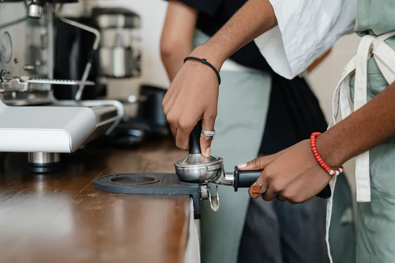 Side view crop faceless ethnic barista wearing apron pressing ground coffee by using tamper before preparing aromatic cappuccino while working in coffeehouse